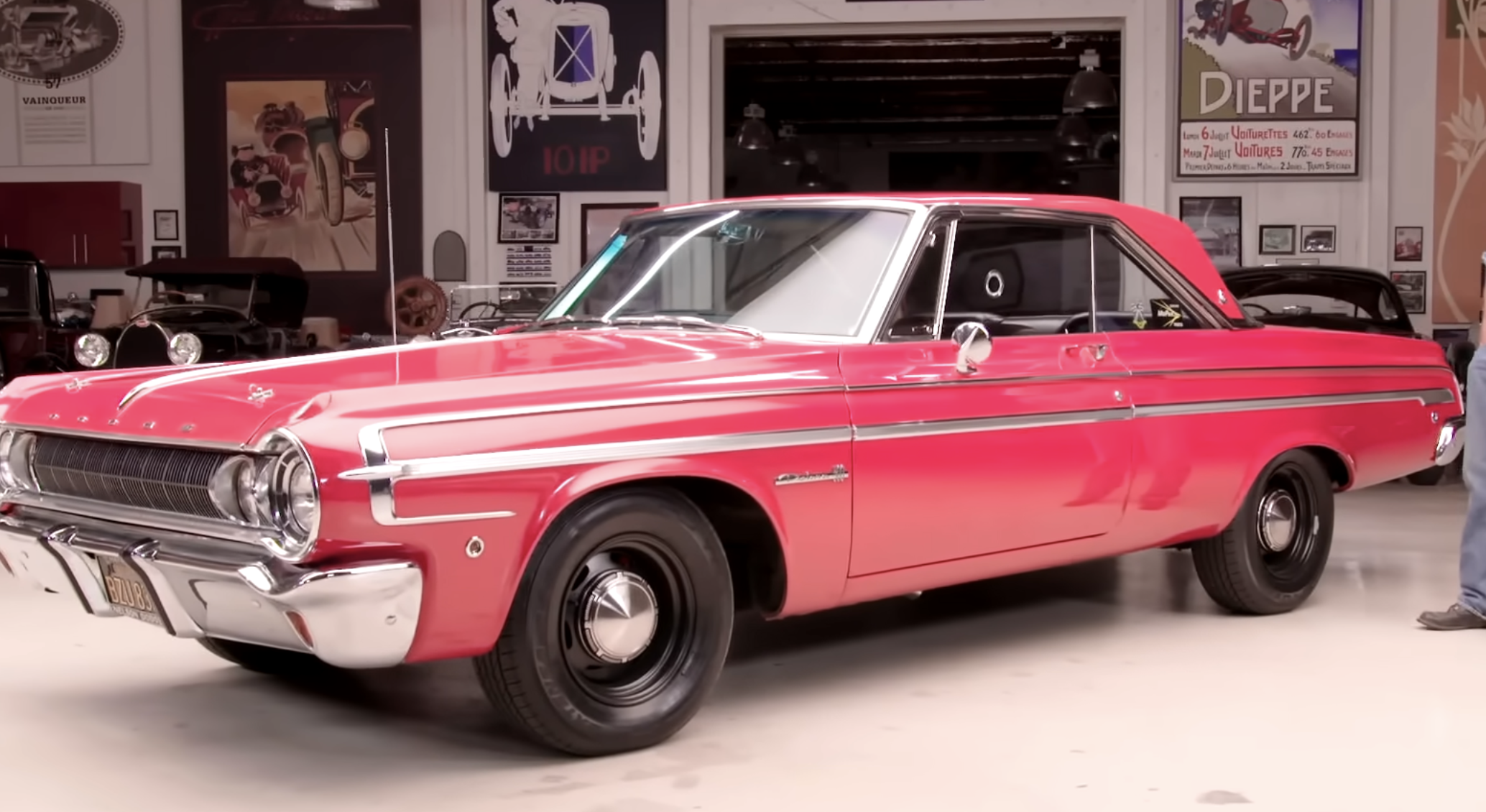 A red vintage Dodge car with chrome detailing is parked indoors in a garage filled with automotive memorabilia and other classic cars.