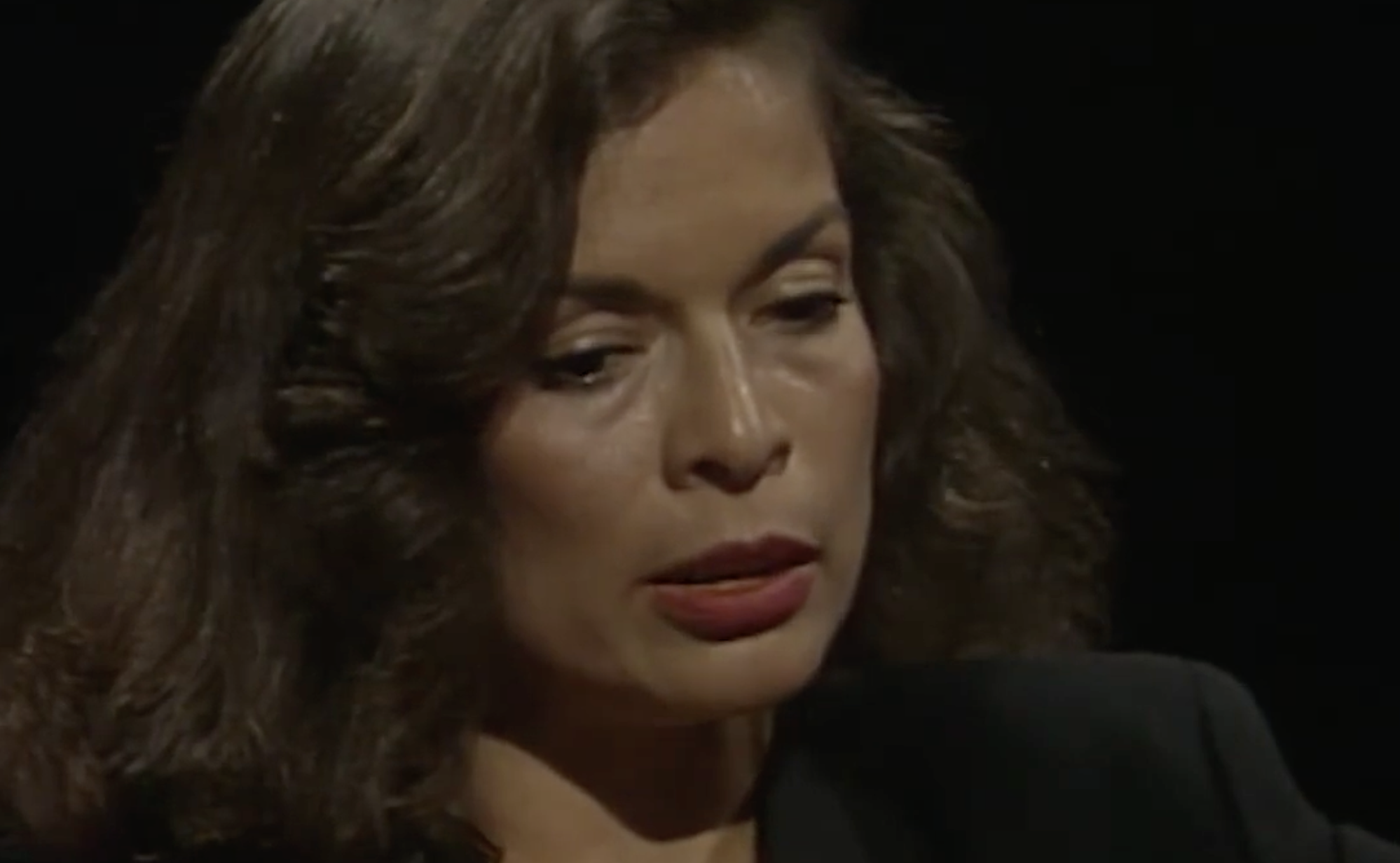 A woman with dark hair looking downward with a thoughtful expression.