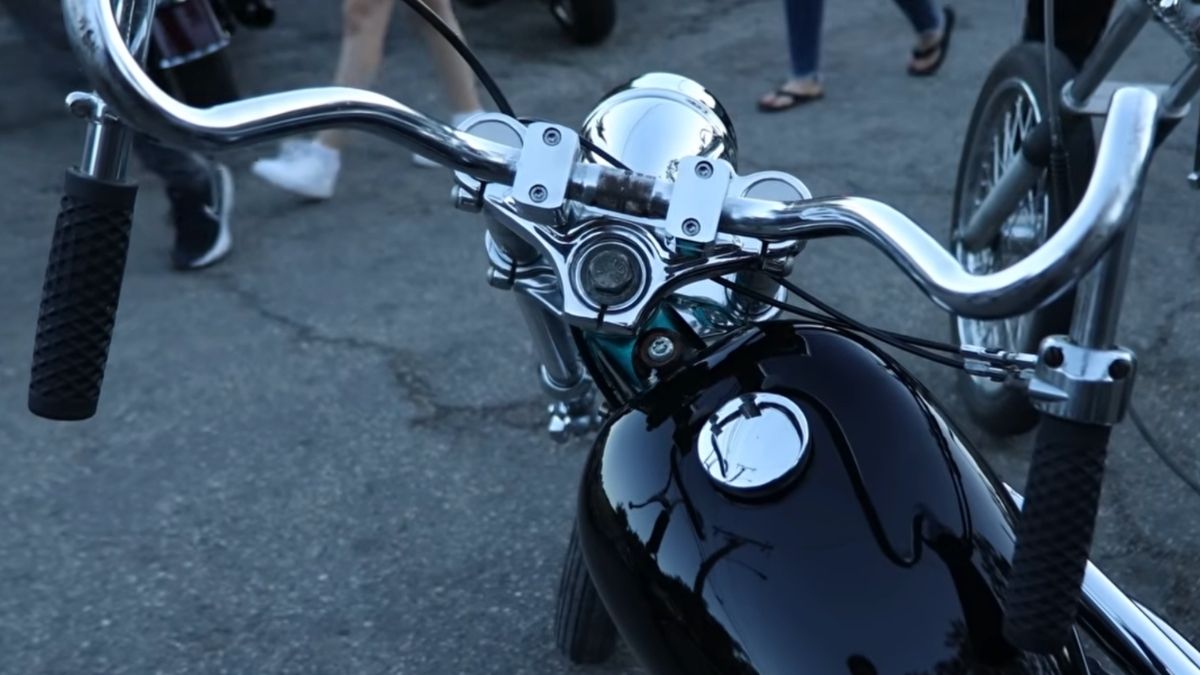 A close-up view of a motorcycle’s handlebars and shiny black fuel tank, with people’s feet visible in the background on asphalt.