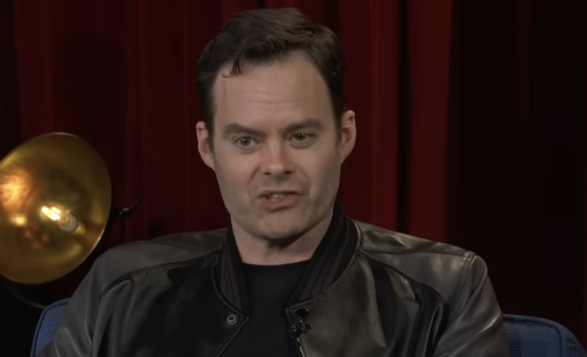 Bill Hader speaking while sitting in front of a red curtain.
