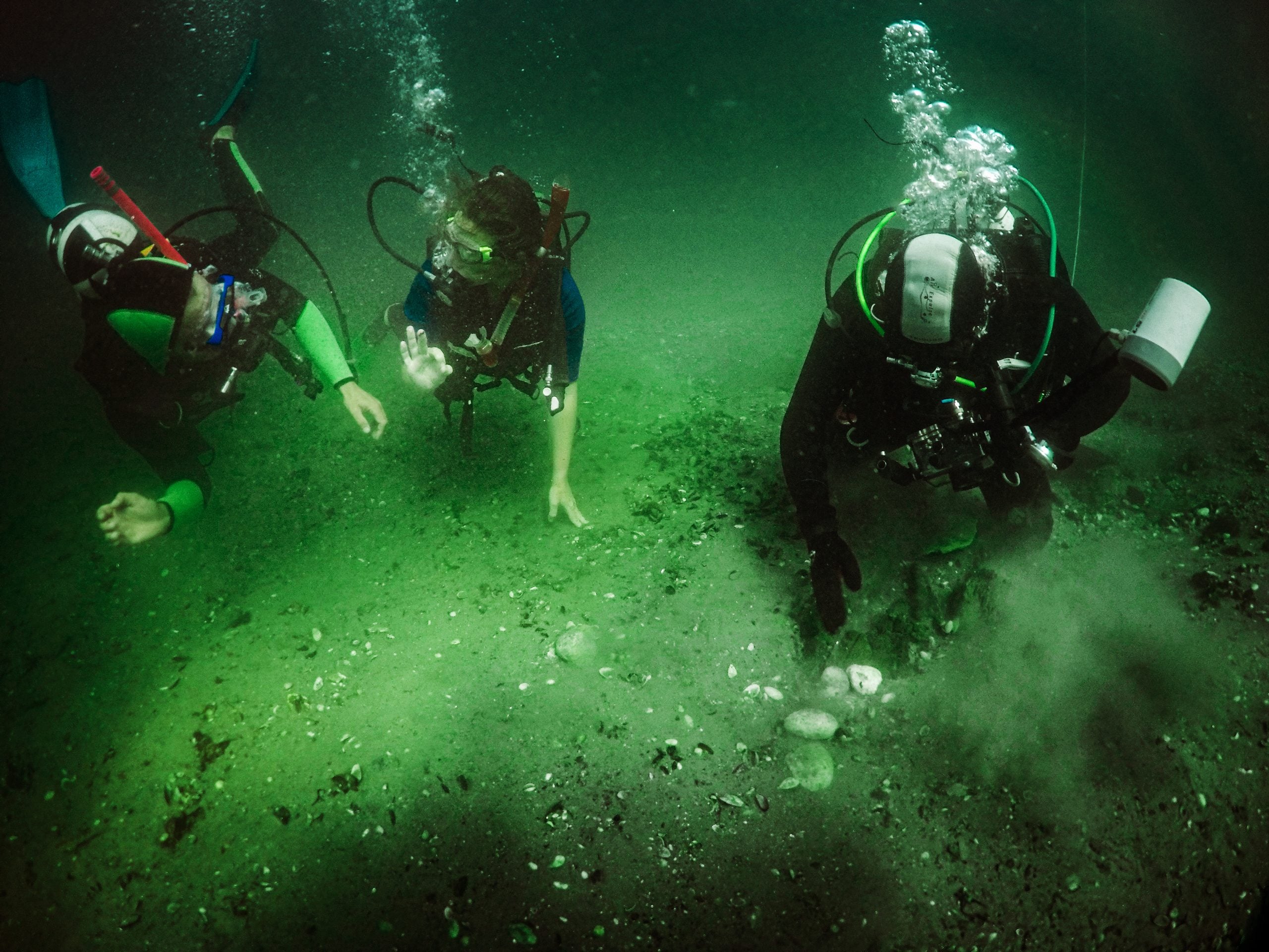 Three scuba divers explore a sandy, murky underwater site, with one diver reaching down to touch the bottom.