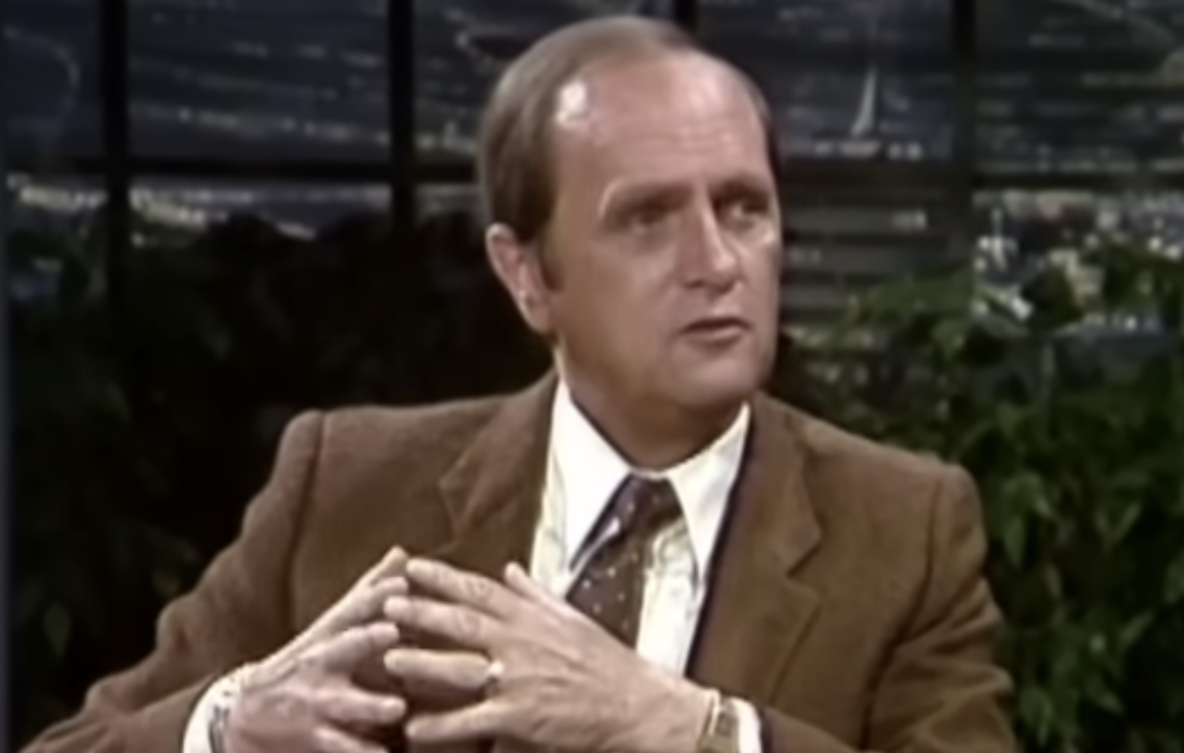 Bob Newhart sitting in an armchair during a televised interview with his hands clasped.