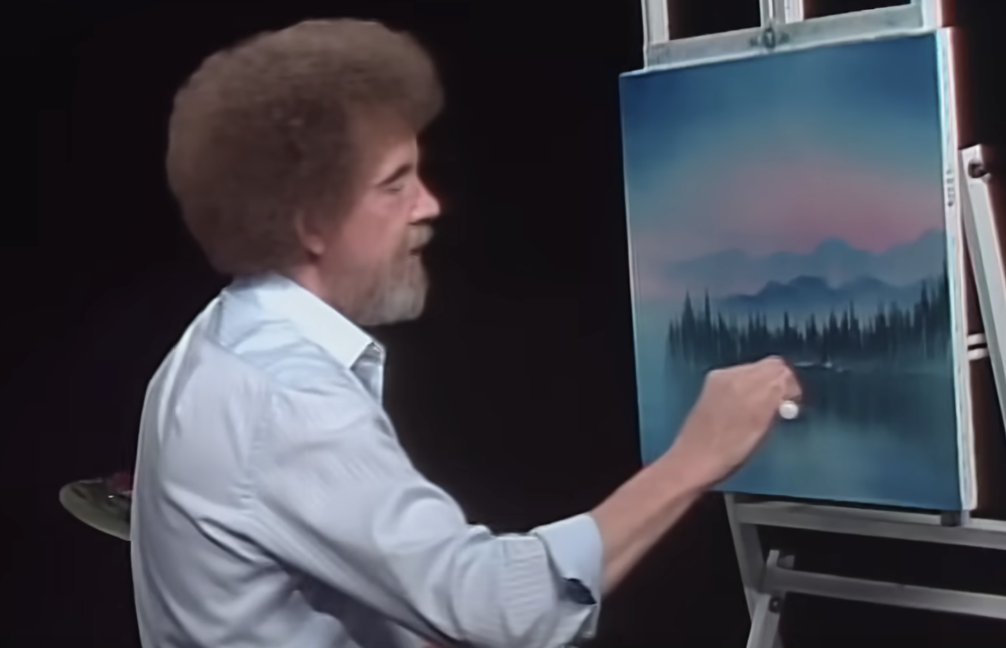 Bob Ross painting a landscape of mountains and trees on a canvas.