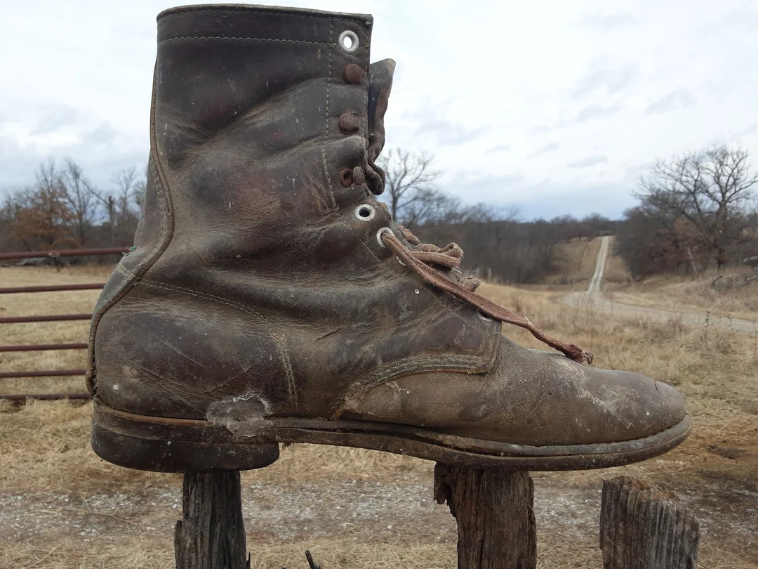 A worn-out brown boot is perched on a wooden fence post in a rural field with a dirt road and trees in the background under a cloudy sky.