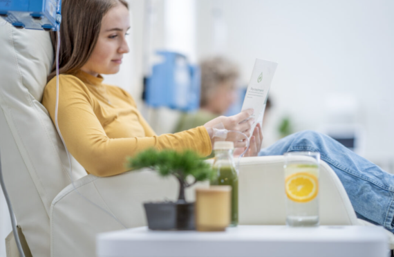 A woman in a yellow shirt sits in a chair receiving intravenous therapy while reading an informational pamphlet.