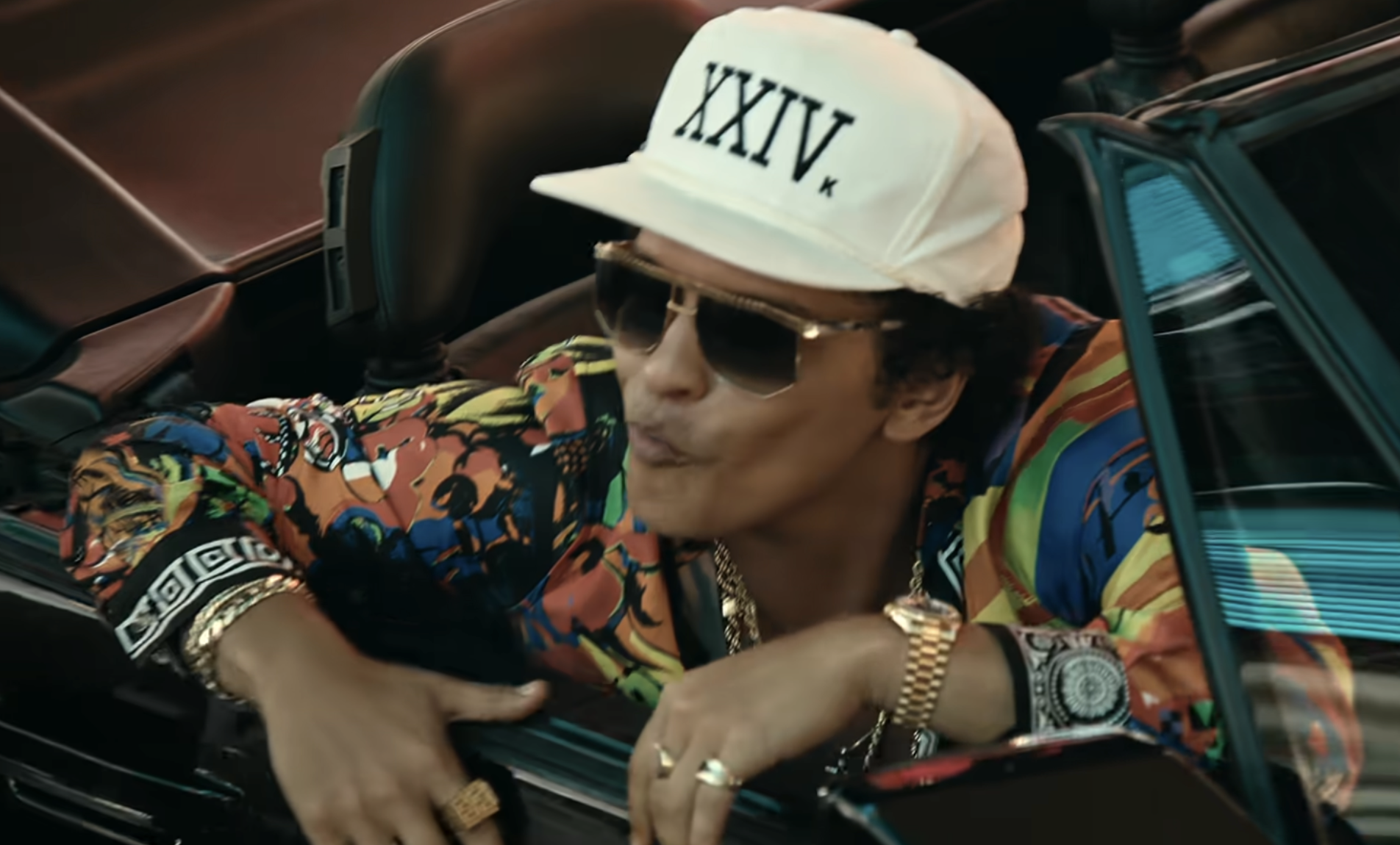 Bruno Mars wearing a white “XXIV” cap and a colorful patterned shirt while leaning out of a convertible.
