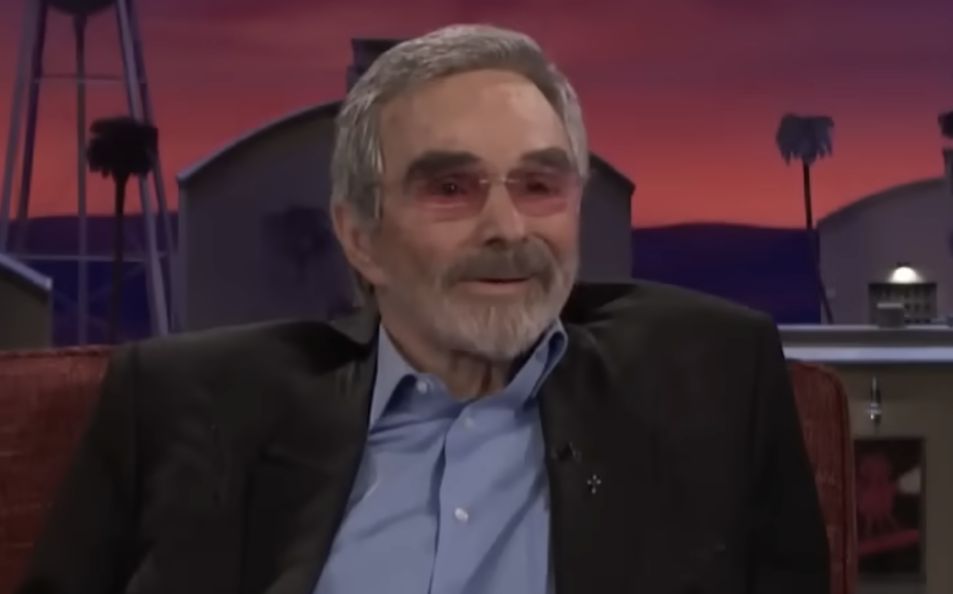 Burt Reynolds with a beard and tinted glasses, wearing a blazer over a button-down shirt during a studio interview.