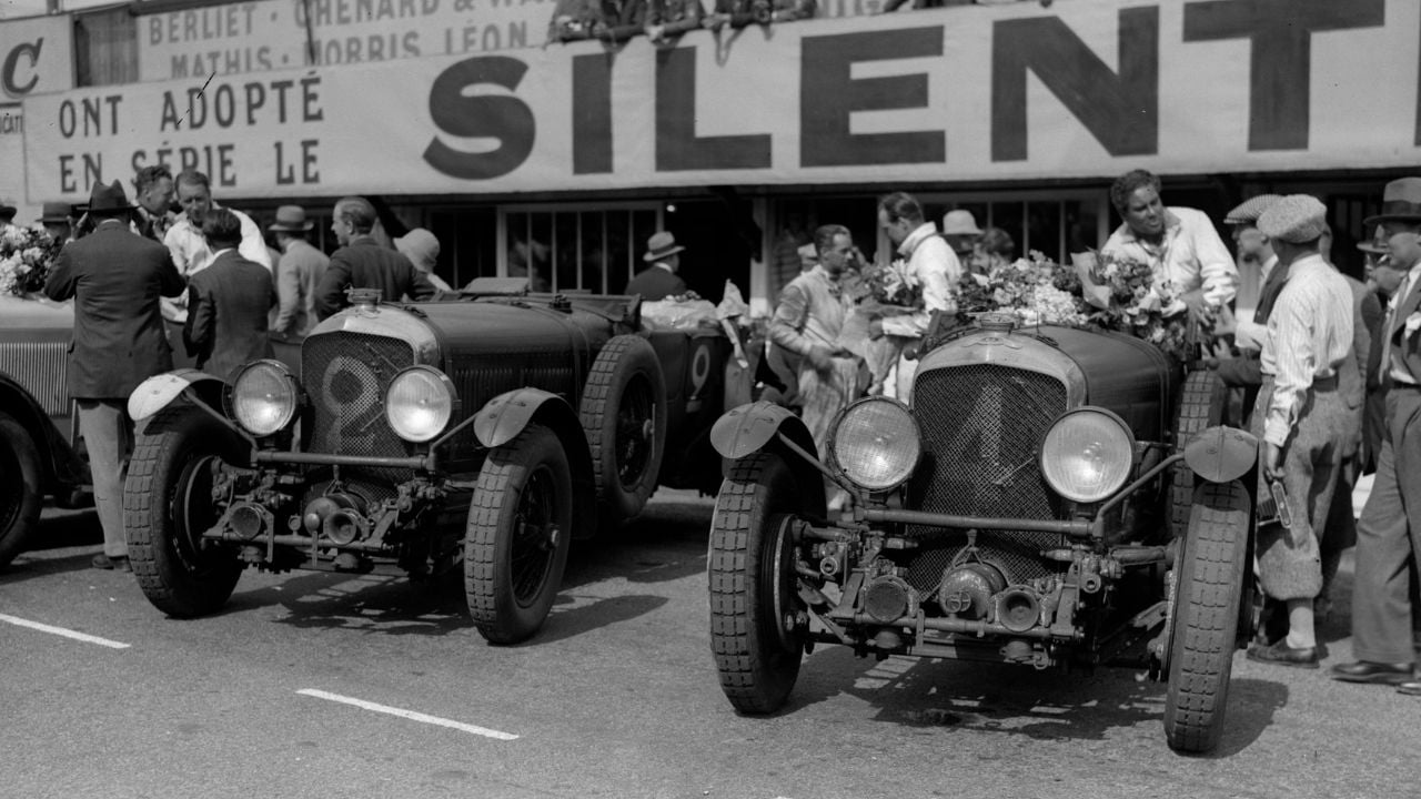 Two vintage race cars with flower wreaths parked in front of a crowd of people. A large banner above reads “SILENT.” The scene appears to be from a historic racing event.