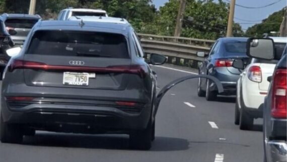 A gray Audi SUV driving on a highway with a broken wheel arch trim panel hanging off its rear tire.