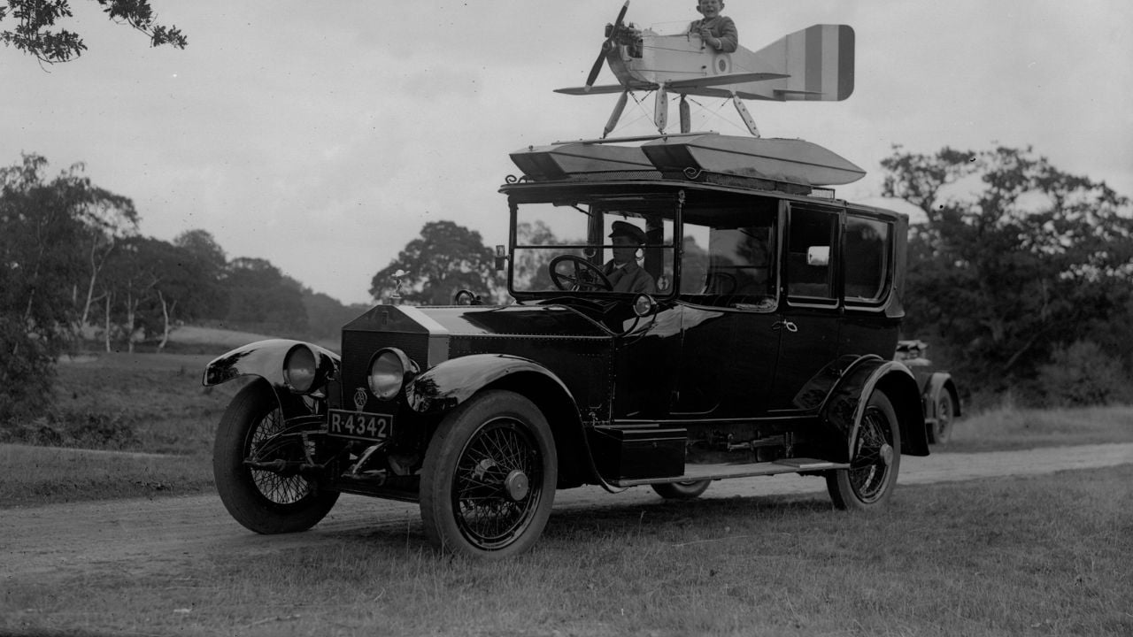 A vintage car with the license plate R-4342 is parked on a dirt road, featuring a small model biplane mounted on its roof. Trees and grassy landscape are visible in the background.