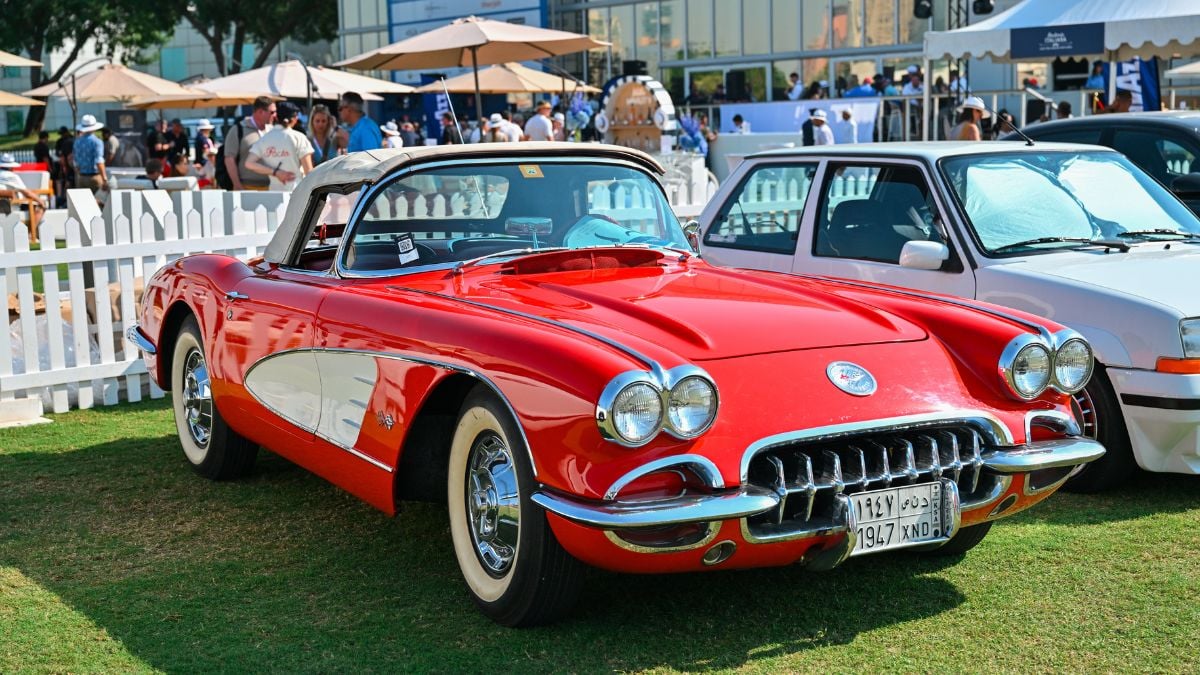 A classic red Chevrolet Corvette convertible is parked on grass at an outdoor car show, next to a white car, with white fencing and people in the background.
