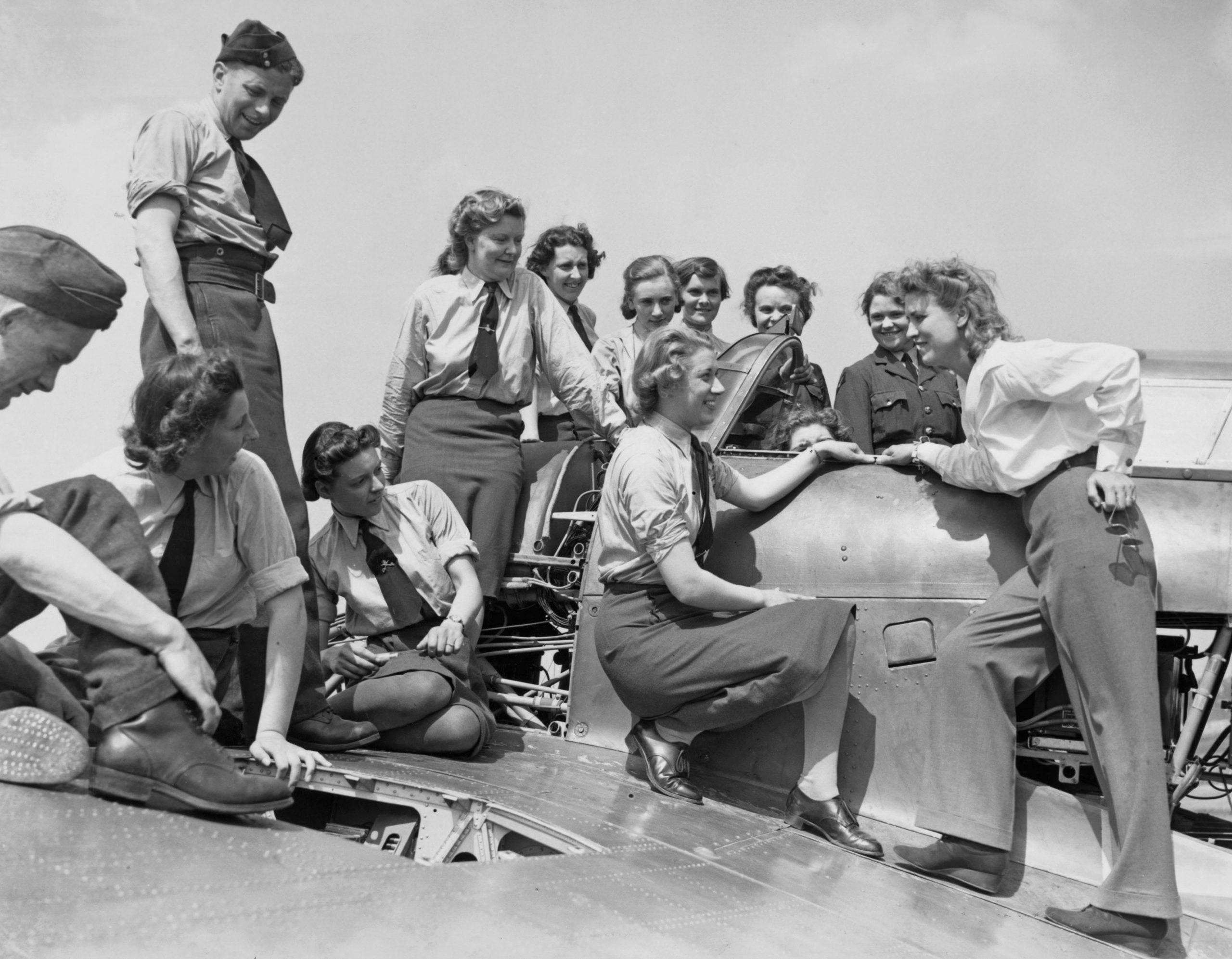 A group of women in uniforms and skirts sit and stand on an aircraft wing, smiling and talking together outdoors on a sunny day.