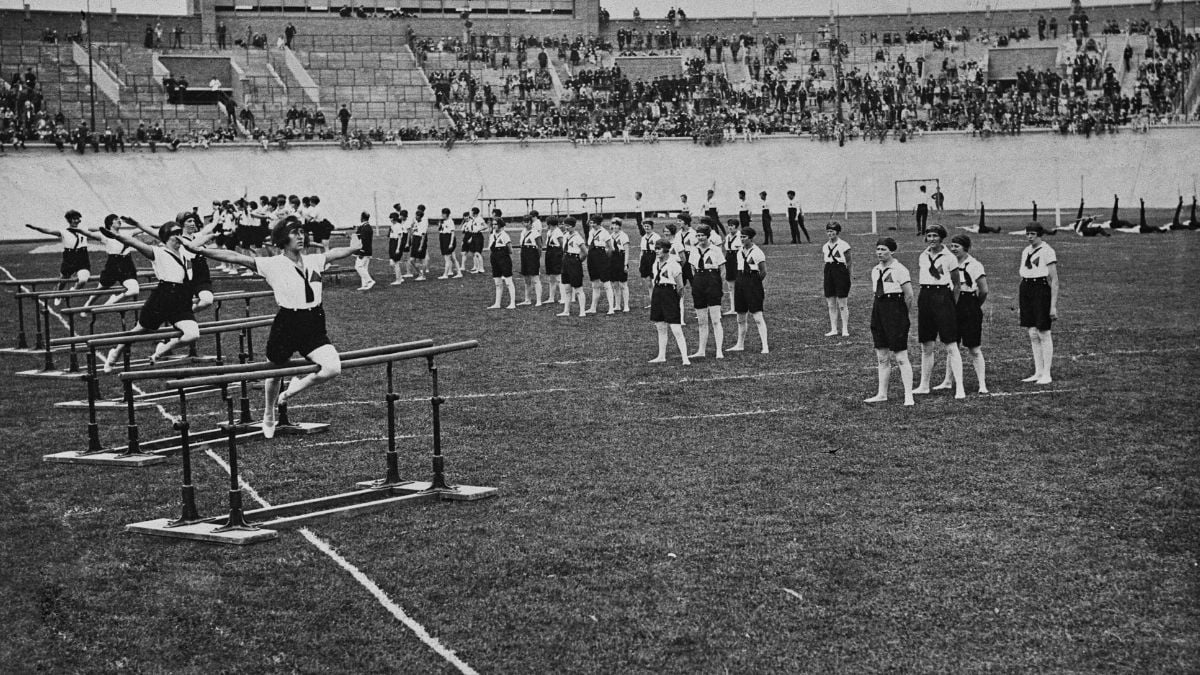 A black-and-white photo shows boys in uniforms doing gymnastics on parallel bars and standing in rows on a field in a stadium with a crowd watching from the stands.
