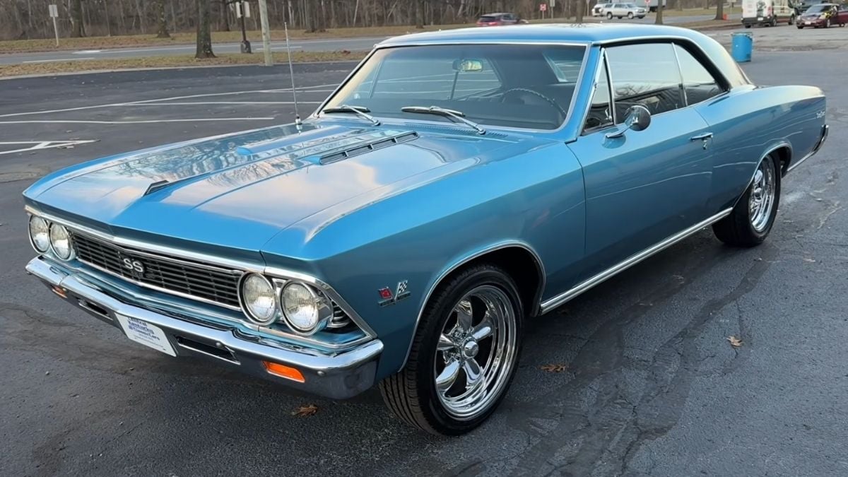 A light blue classic car, likely a 1960s Chevrolet Chevelle SS, is parked in a mostly empty lot. The car has chrome wheels, a white roof, and shiny bumpers, with trees and a distant road in the background.