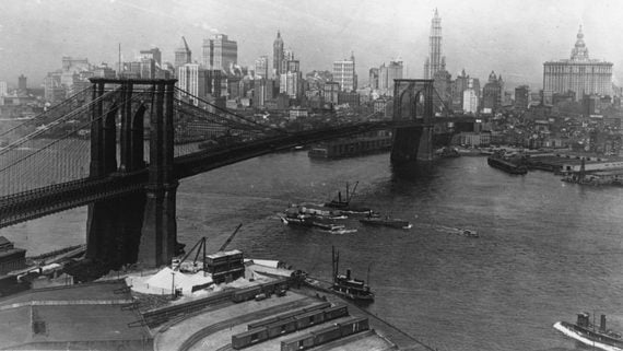 Black and white photo of the Brooklyn Bridge over the East River with several boats below. The New York City skyline with historic skyscrapers is visible in the background.