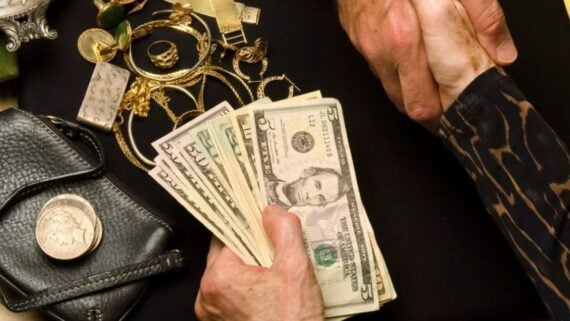 A person holds a fan of US dollar bills over a table scattered with gold jewelry and a leather wallet.