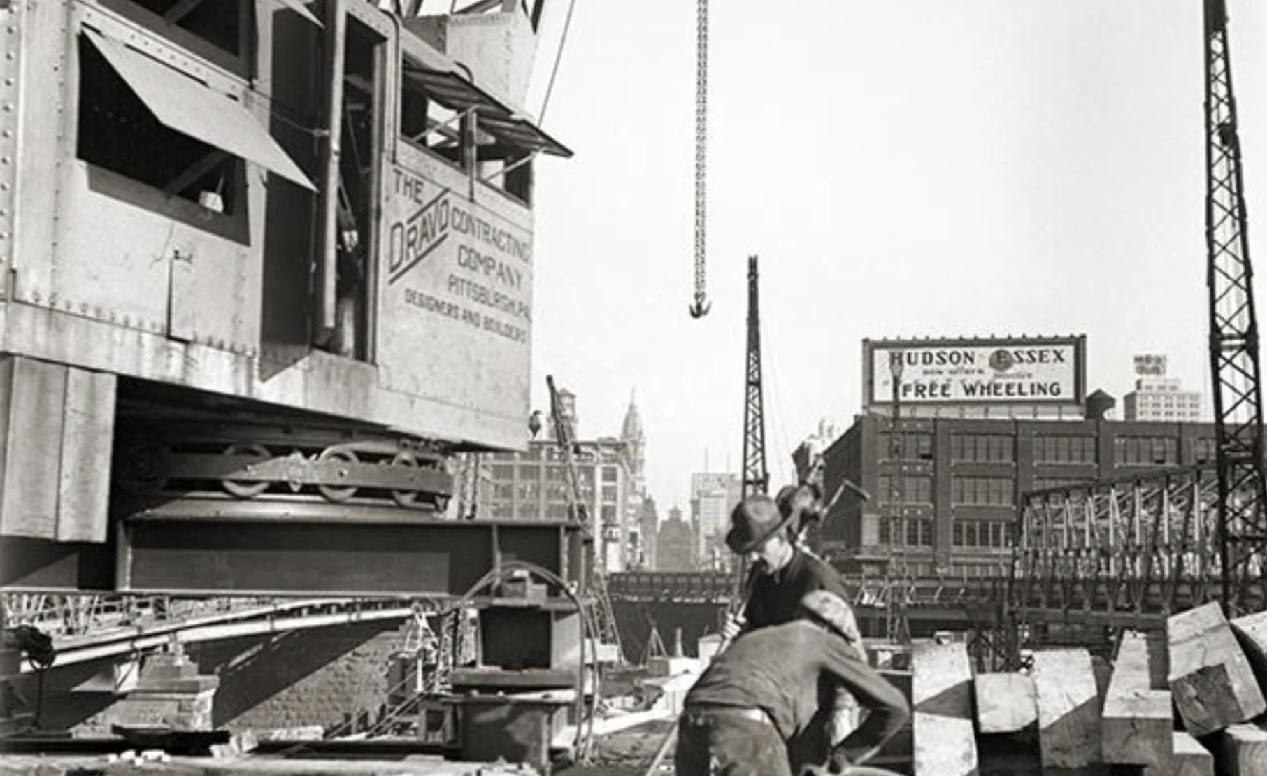 Workers operate at a construction site with a large crane and a billboard advertising Hudson Essex cars in the background.
