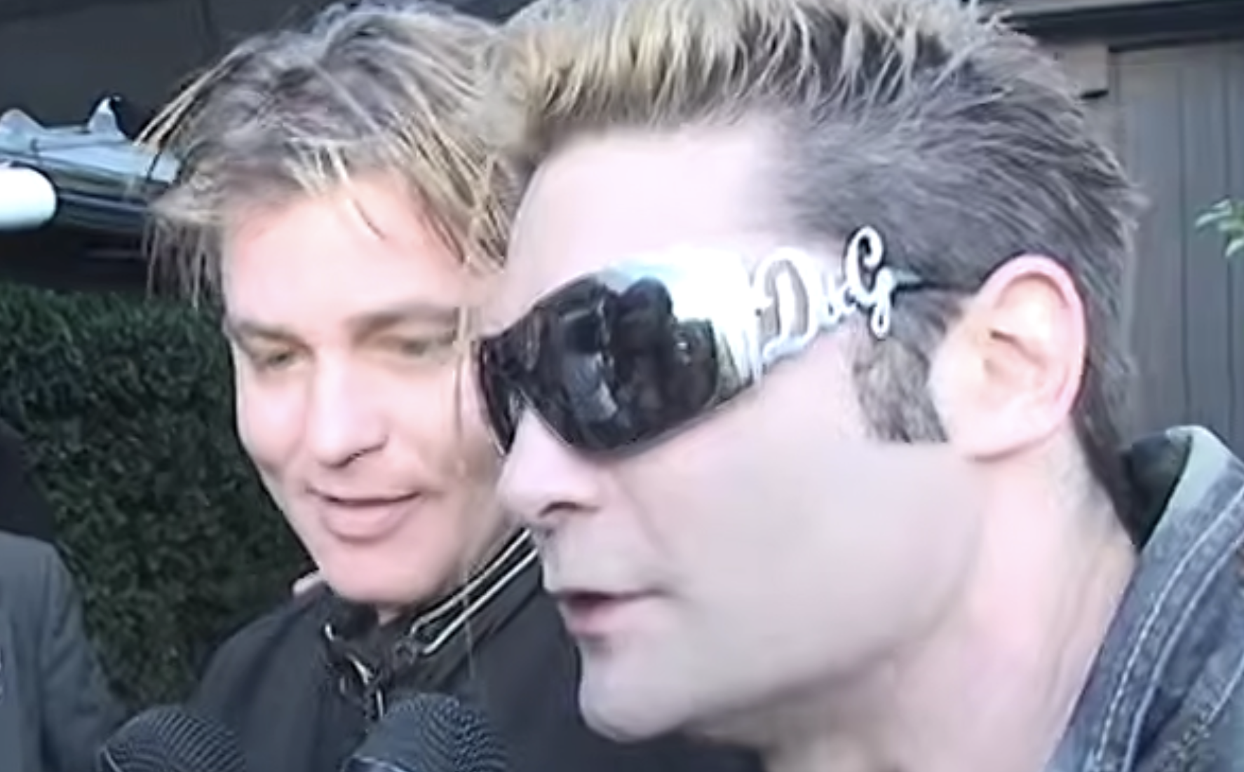 Two men side-by-side, one wearing wraparound sunglasses with prominent silver lettering on the temple.