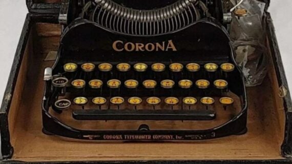 A vintage black Corona portable typewriter with yellow keys sits inside its open carrying case.