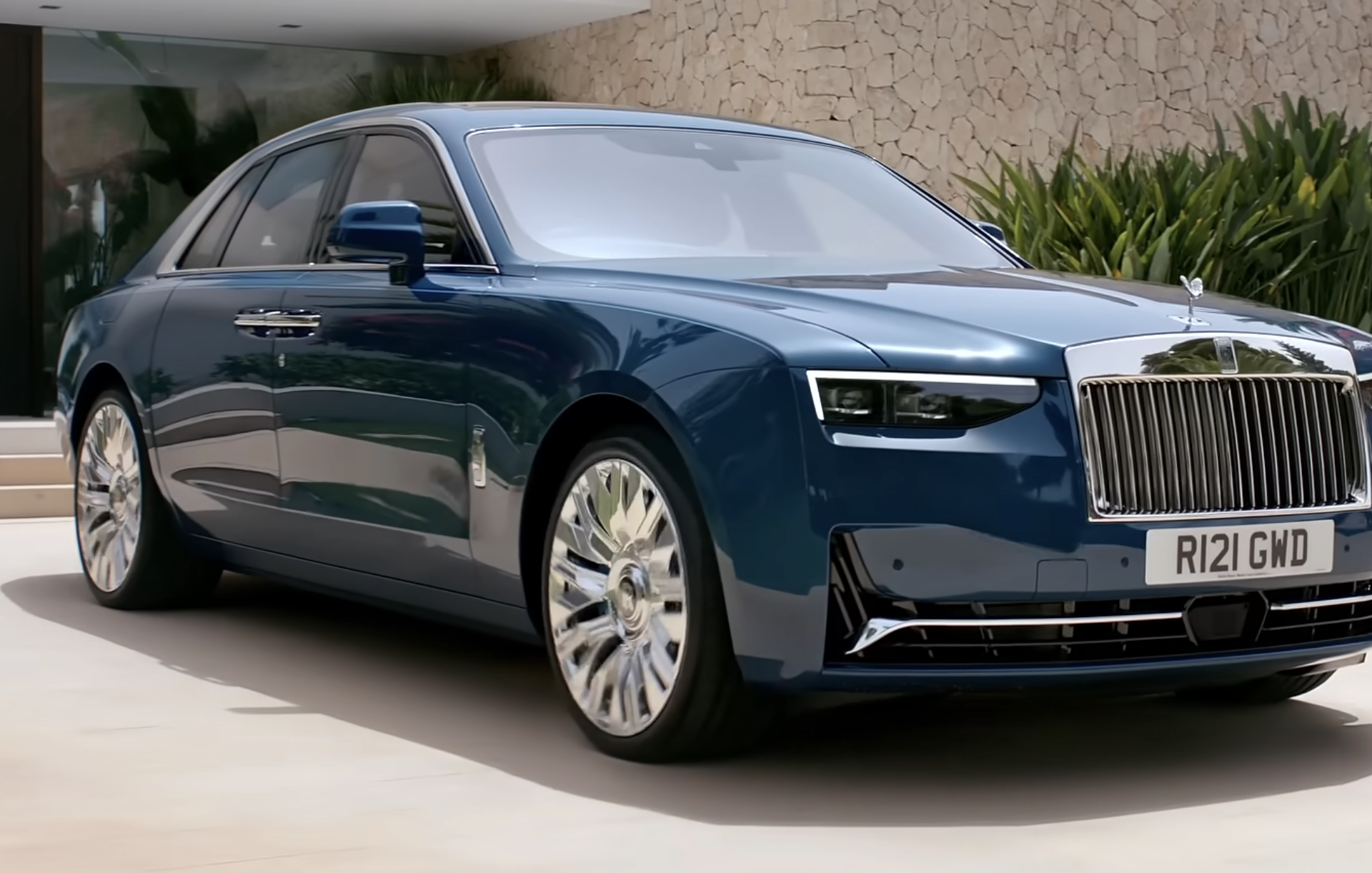 A dark blue Rolls-Royce sedan parked on a paved driveway in front of a stone wall.