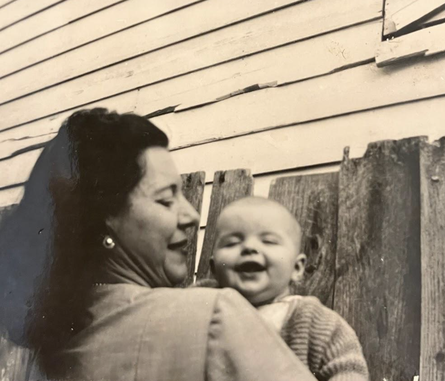 A woman in profile holds a laughing baby in front of a wooden fence and house siding.