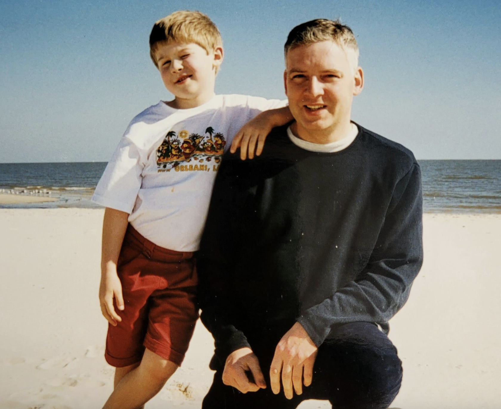 A boy standing on a beach with his arm around a man who is kneeling beside him.