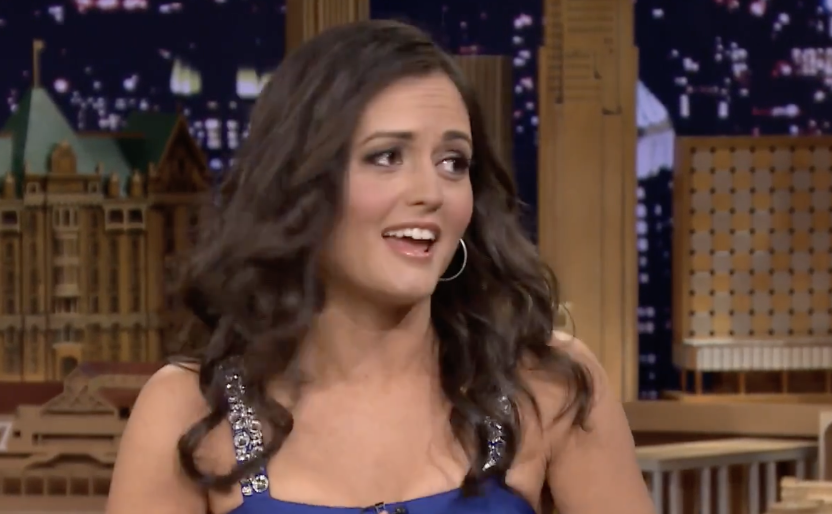 Danica McKellar sitting on a talk show set with a city skyline background.