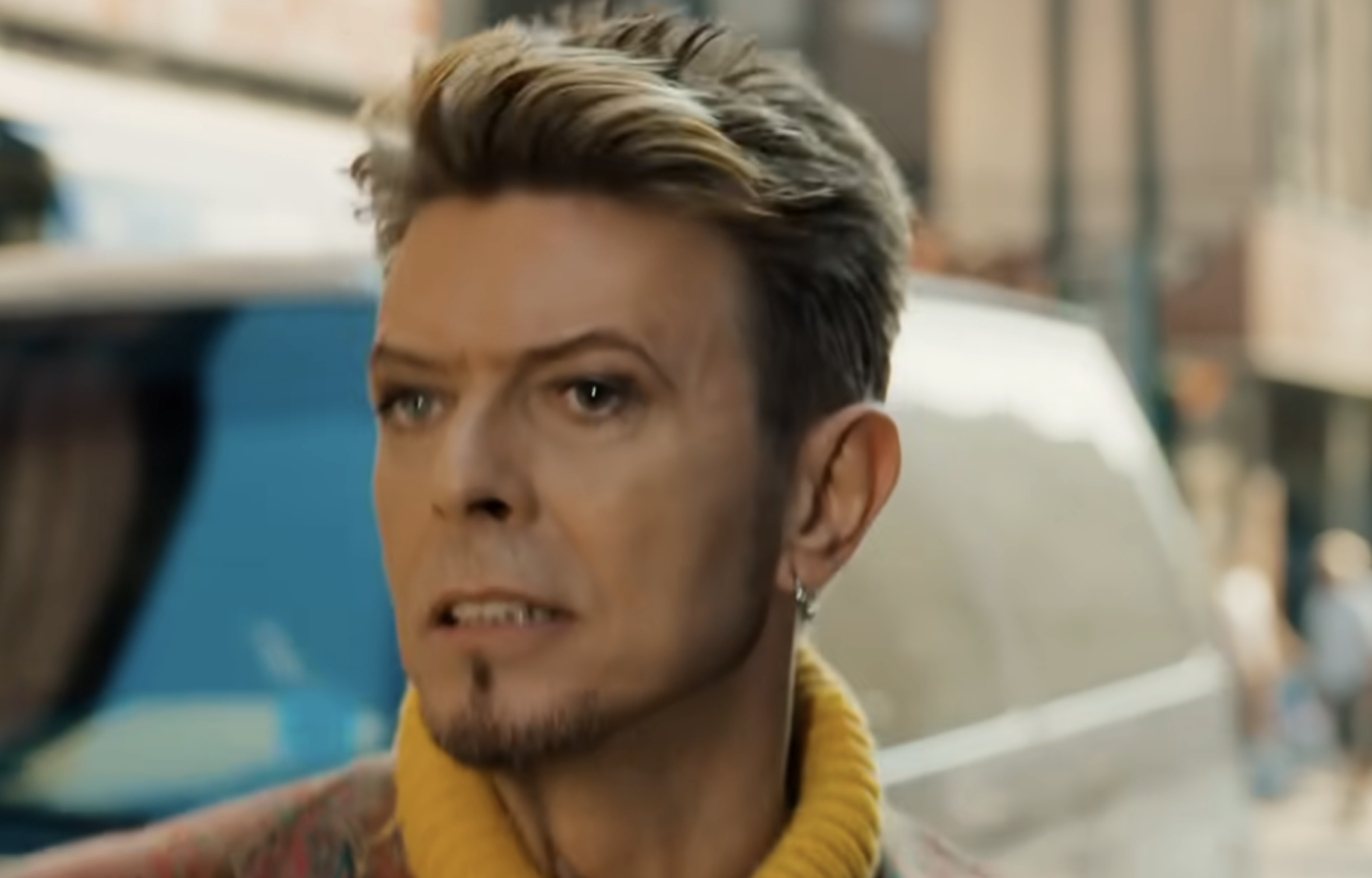 David Bowie with short blonde hair and a goatee, wearing a yellow turtleneck, looking off-camera.