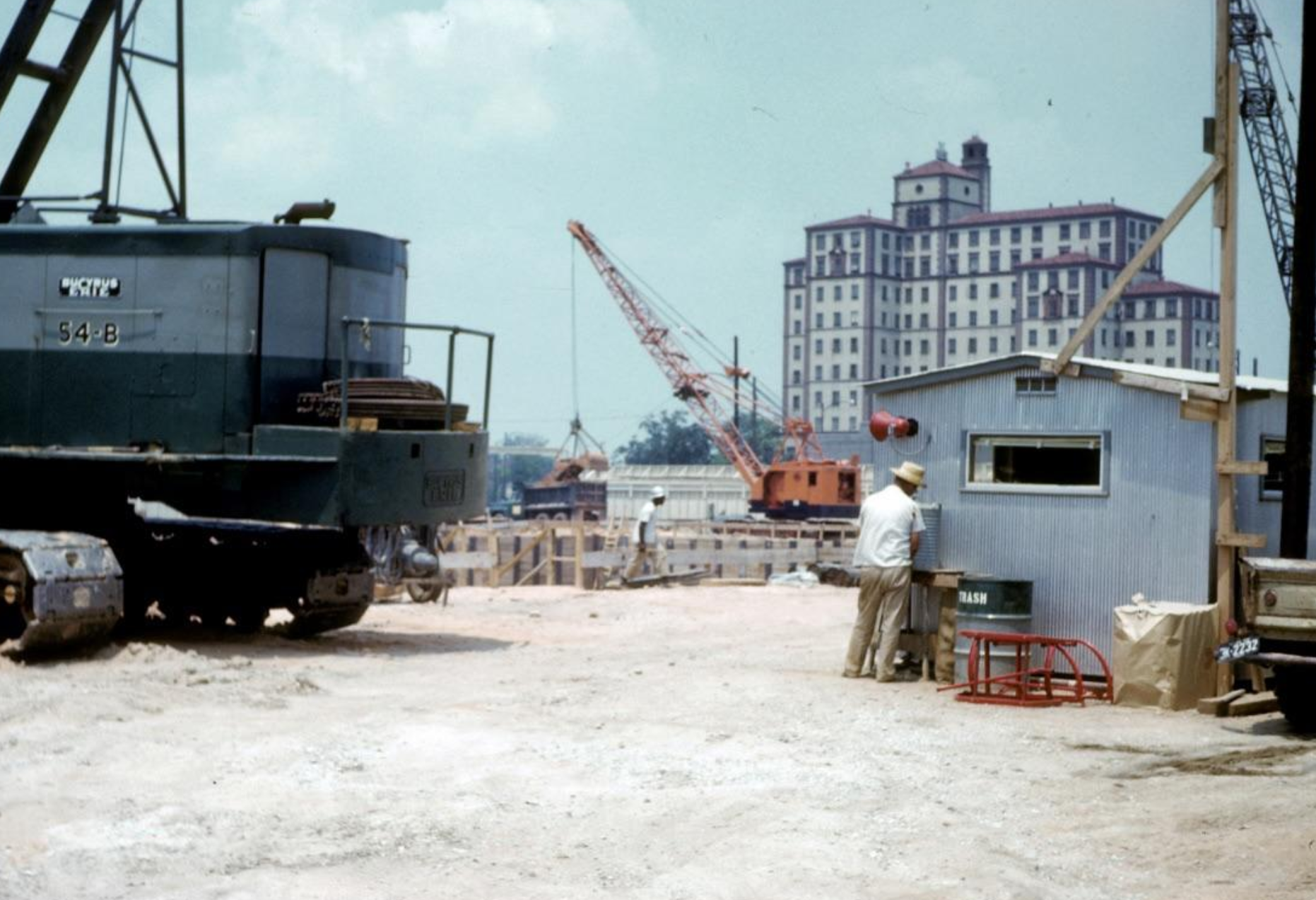 A construction site with two large cranes, a worker standing outside a small metal office, and a large multi-story building in the background.