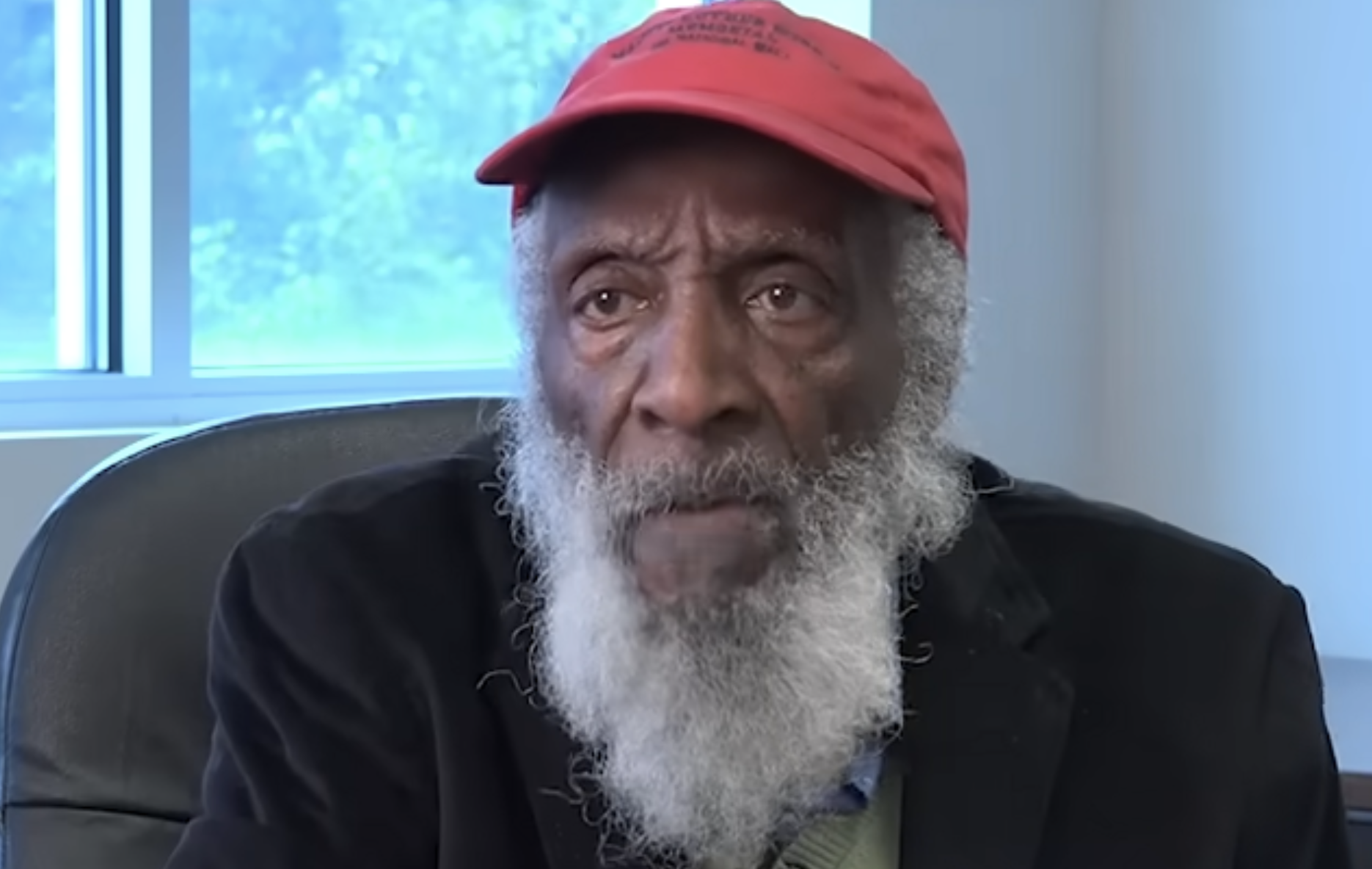 An elderly Black man with a long white beard wearing a red baseball cap and a dark blazer.