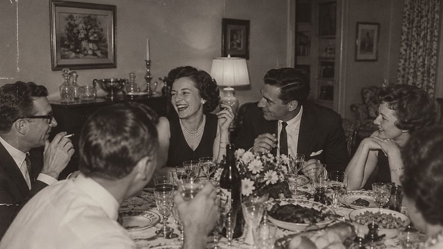 A group of people laughing and talking while sitting at a dinner table set with wine glasses and food.