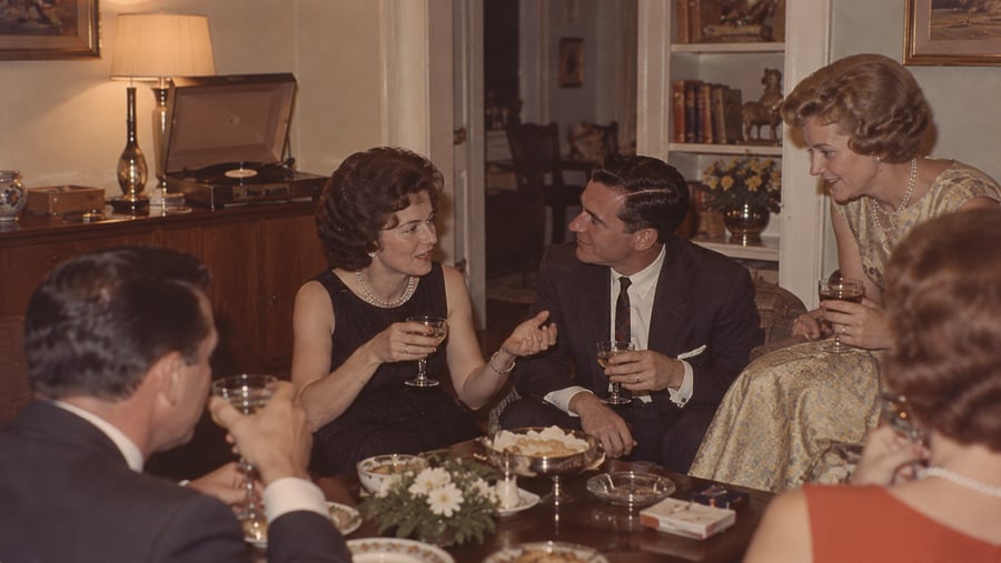 Four people sit around a low table with drinks and snacks in a living room, engaged in conversation.