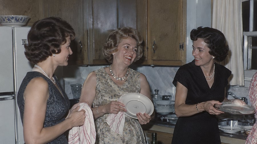 Three women in vintage dresses and pearl necklaces stand in a kitchen, talking and holding plates.