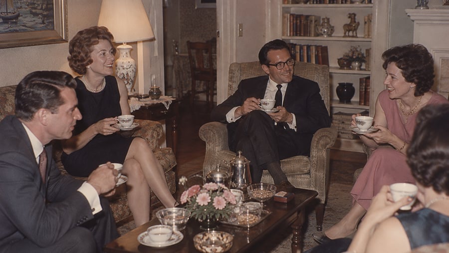 Two couples sit in a living room, drinking tea and conversing around a coffee table with a flower arrangement and service set.