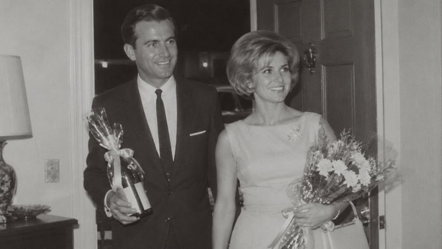 A smiling man in a suit holding a gift-wrapped bottle and a smiling woman in a dress holding a bouquet of flowers standing in a doorway.
