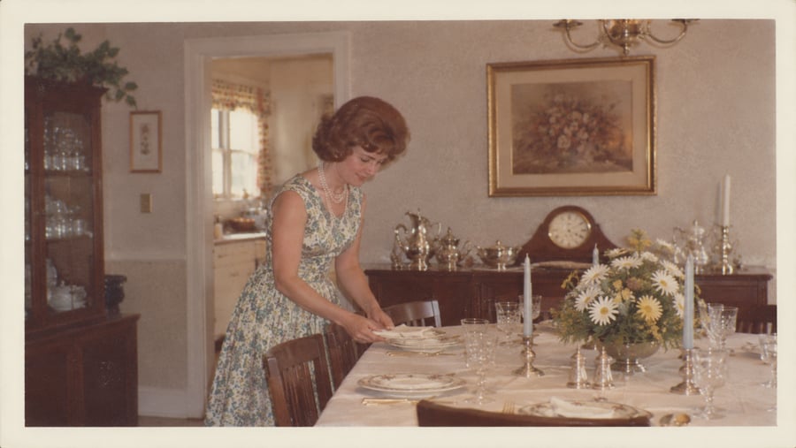 A woman in a floral dress sets a formal dining table.