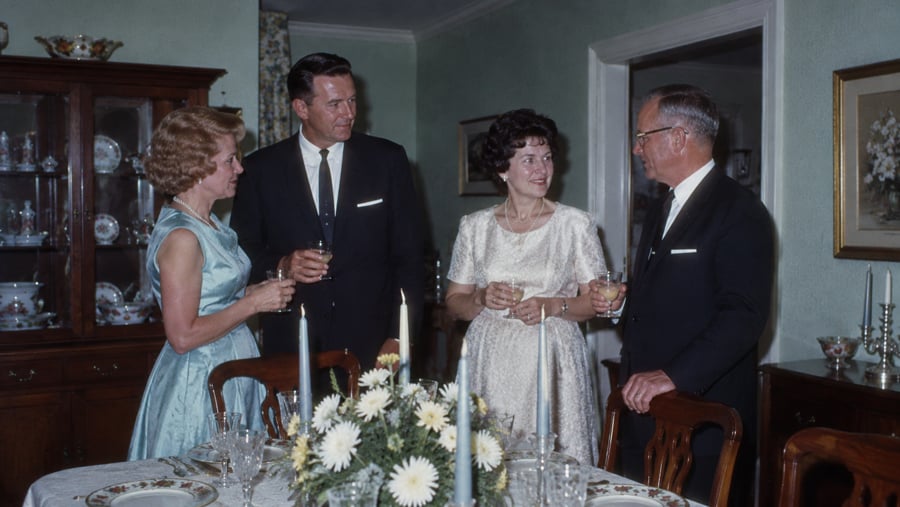Two couples standing around a dining table holding drinks and talking.