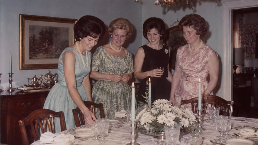 Four women in formal dresses standing around a dining table set for a meal.