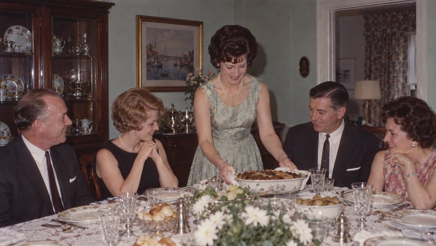 A woman in a floral dress serves a casserole to three people seated at a dining table set with china and floral centerpieces.