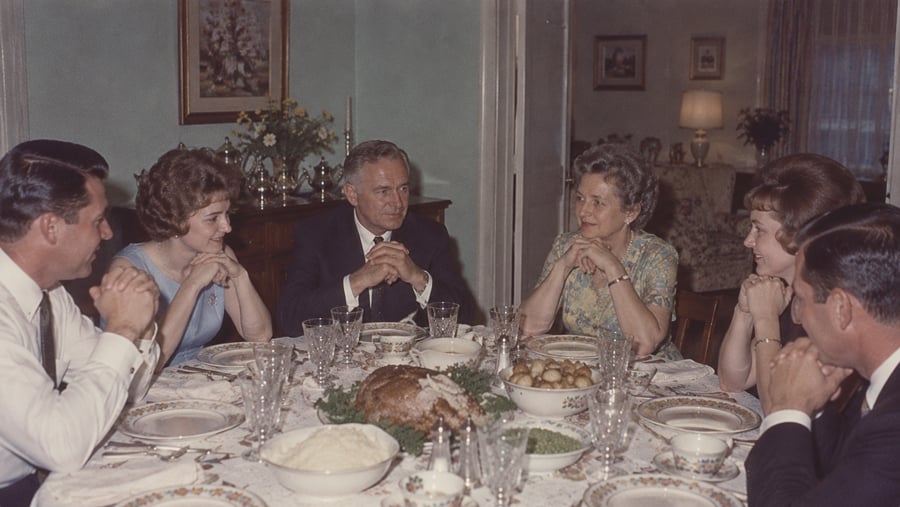 Six adults seated around a dining table set for a formal meal with a roasted bird at the center.