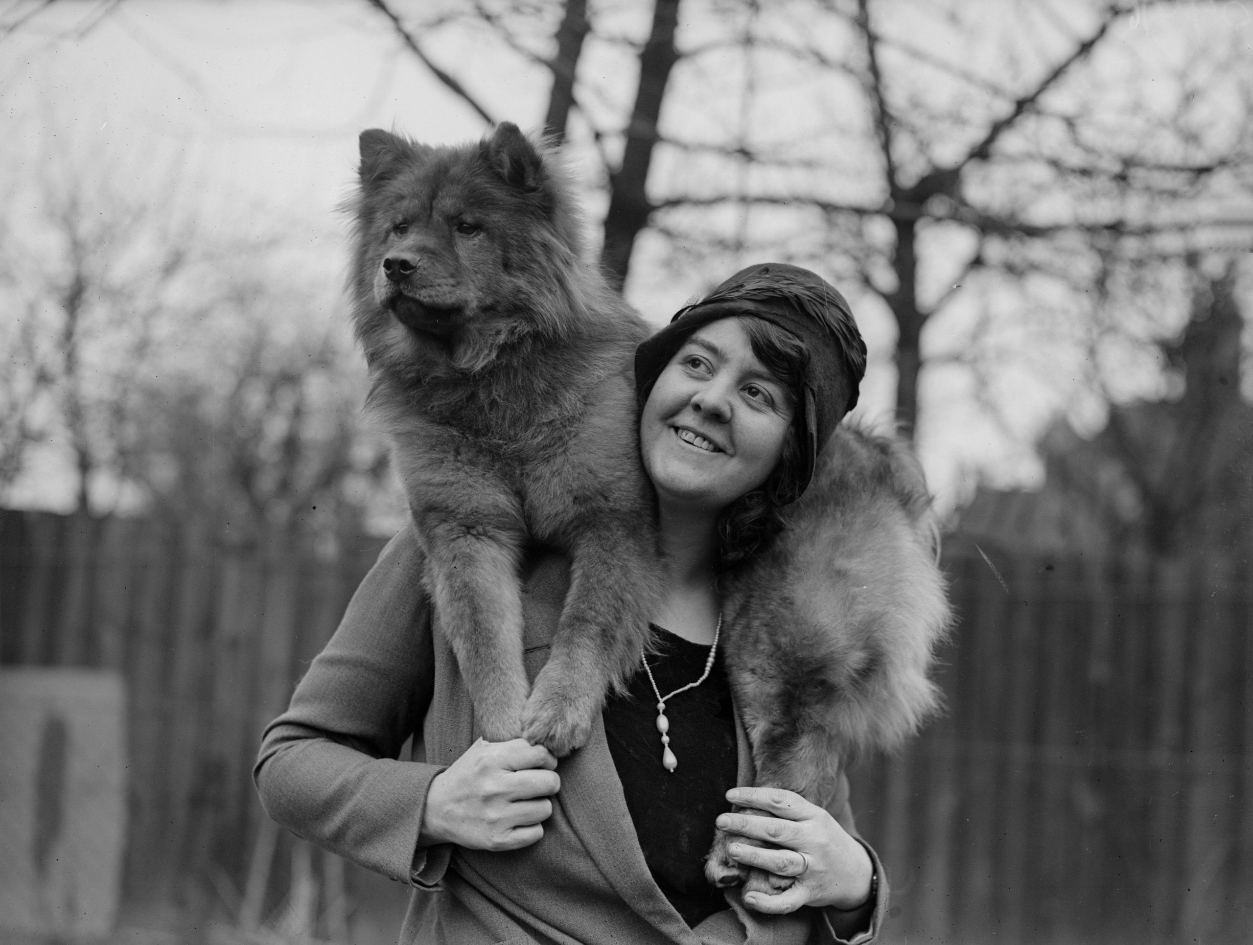 A smiling woman stands outdoors with a fluffy dog draped over her shoulders. Leafless trees and a wooden fence are visible in the background.