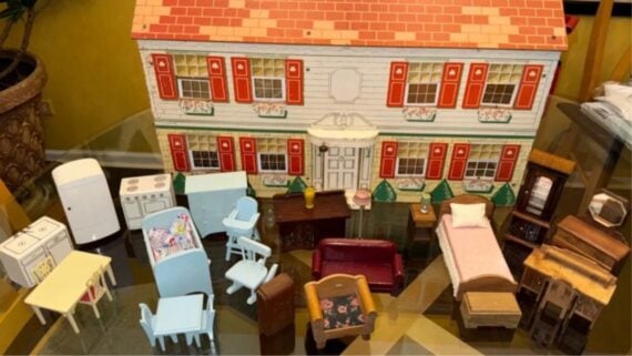 A vintage metal dollhouse with a two-story design, displayed with an assortment of miniature toy furniture including beds, chairs, tables, and kitchen appliances.