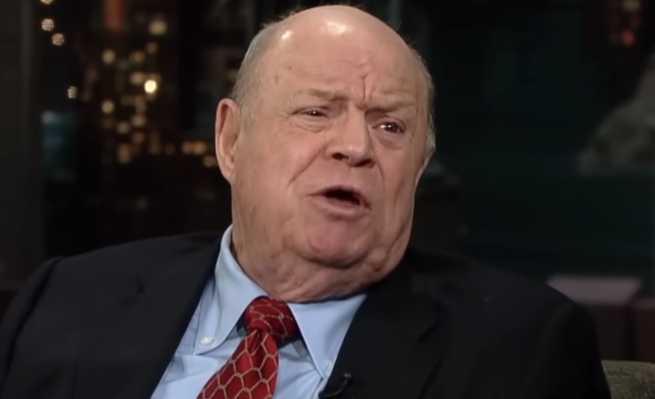 Don Rickles wearing a dark suit, light blue dress shirt, and red patterned tie.