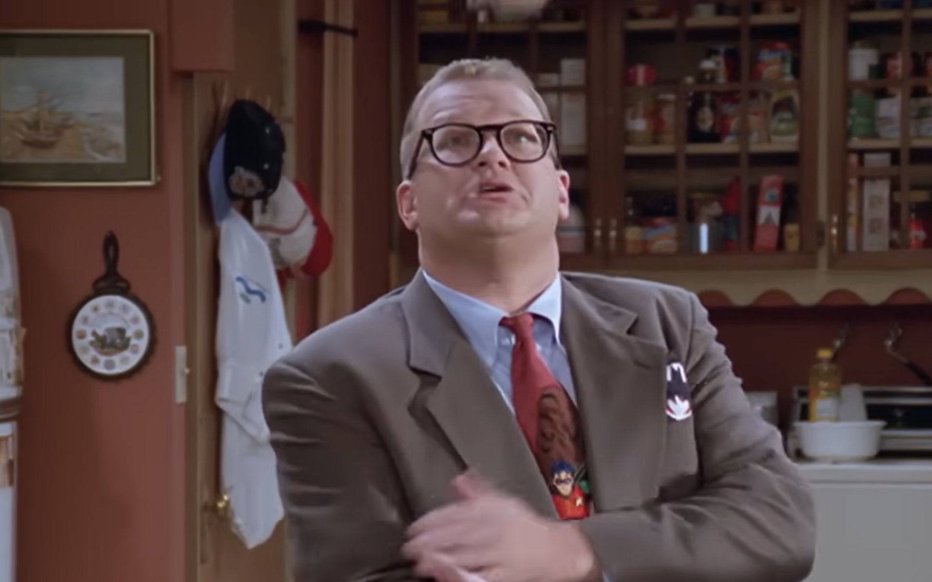 Drew Carey in a brown suit and red tie looking upward in a kitchen.