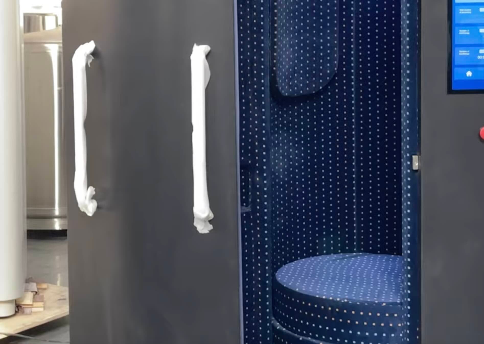 A tall, dark gray privacy booth with a partially open door, revealing a blue, polka-dotted interior with a curved bench and a digital touchscreen on the exterior.