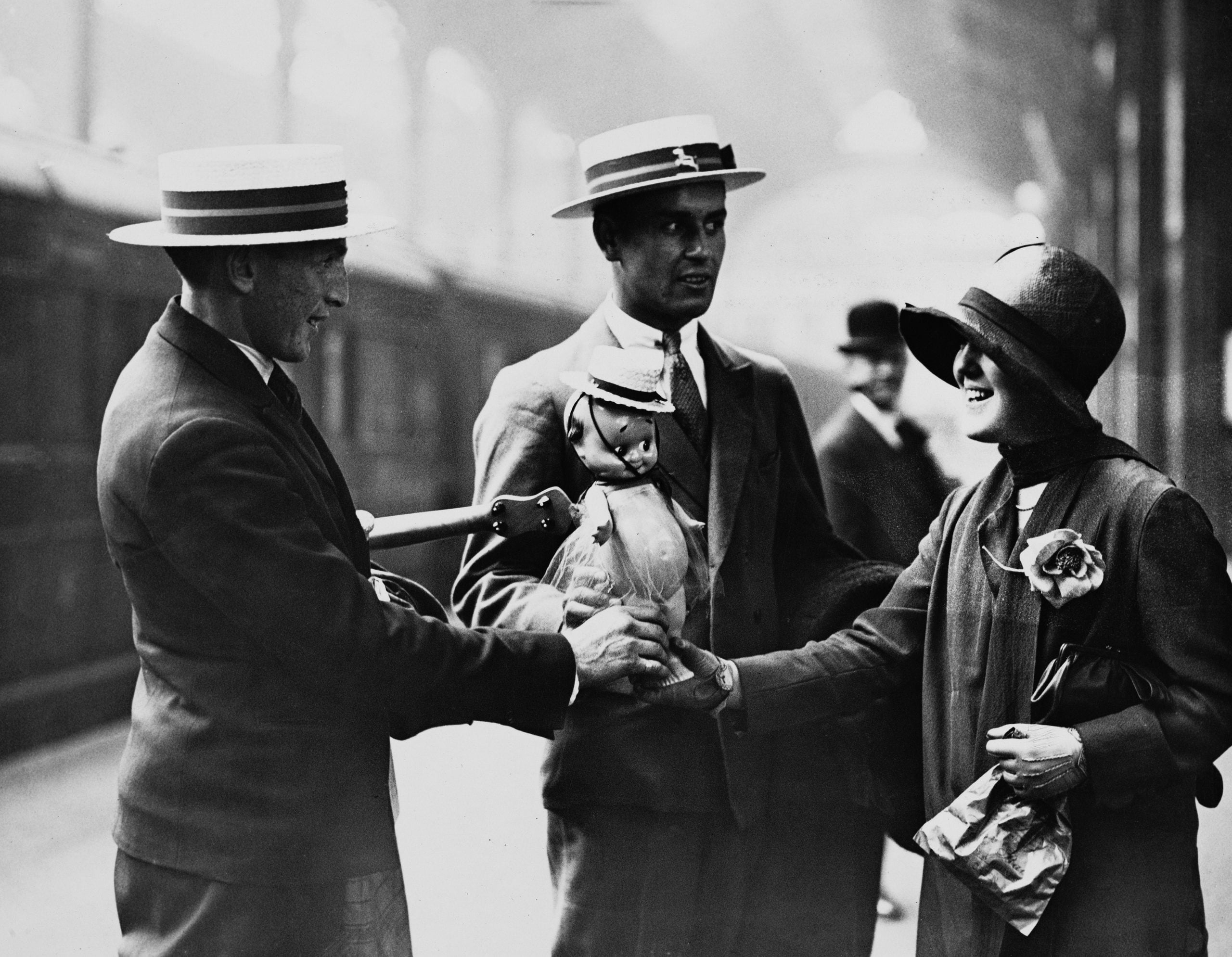Three people in vintage clothing and hats greet each other at a train station. One man holds a small dog dressed in a coat, while the woman smilingly shakes hands with another man.