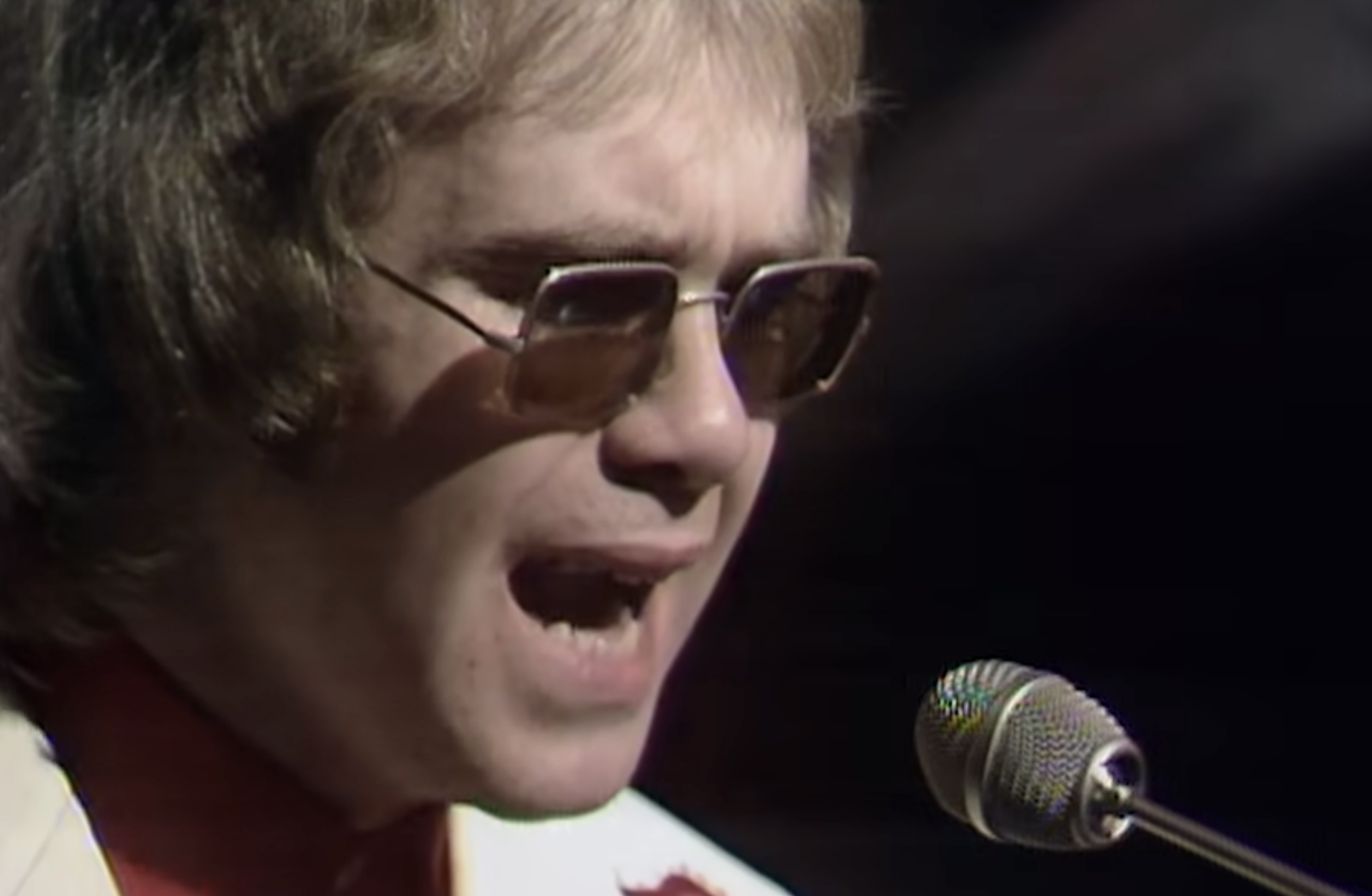 Elton John singing into a microphone while wearing tinted glasses.