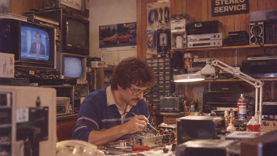 A man with a mustache and glasses works at a cluttered electronics repair bench surrounded by vintage televisions and stereo equipment.