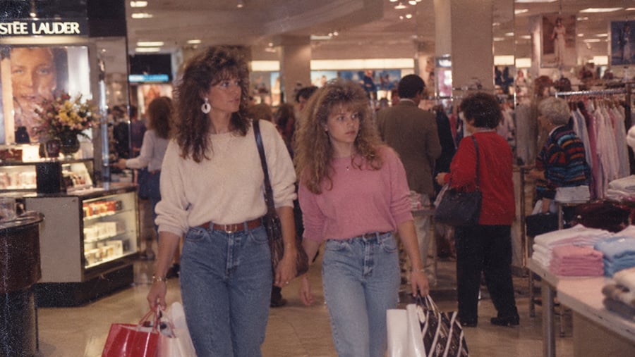 Two women with voluminous hair walking through a department store while carrying shopping bags.