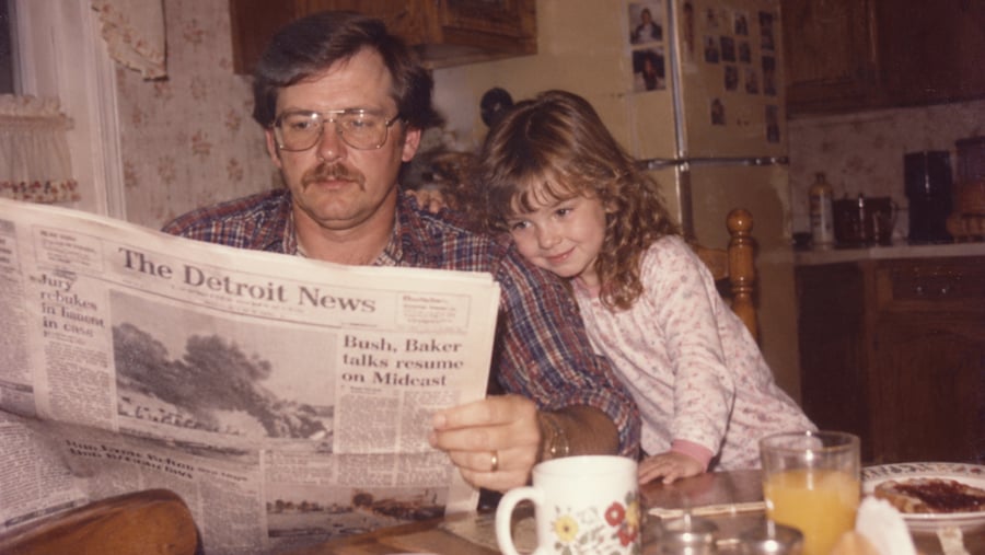 A man in glasses reading a Detroit News newspaper at a kitchen table while a young girl leans against his arm.